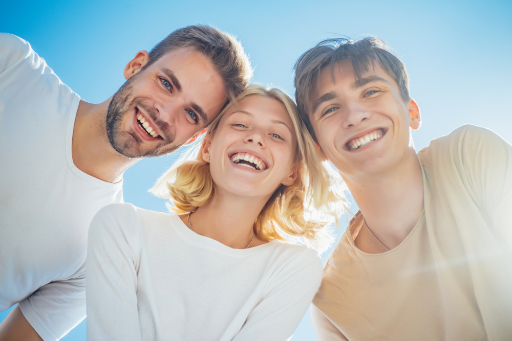 Group of happy friends smiling under the sunlight – Dentist Aliso Viejo Three cheerful young adults smiling and standing together outdoors under bright blue sky – Dentist Aliso Viejo